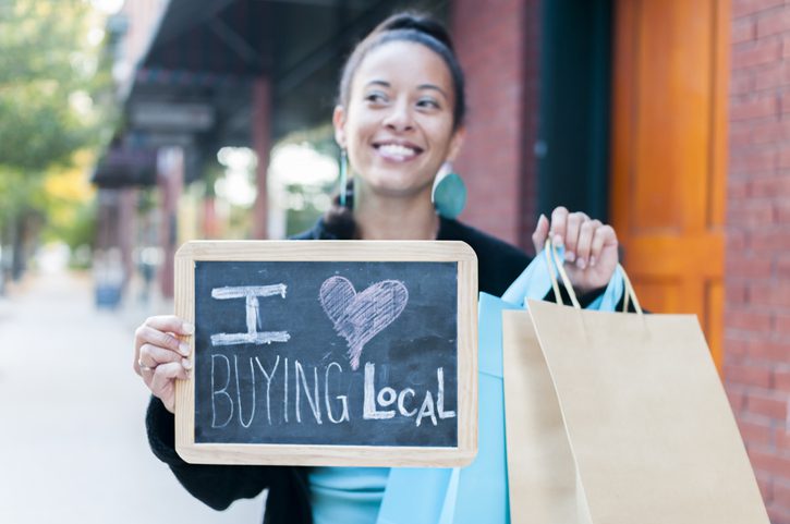 Woman with shopping bags holding up a "I Love Buying Local" sign on a local downtown street.