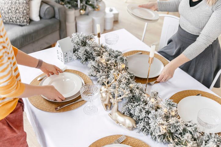 Holiday table decorated with golden accents, white tablecloth and flocked pine tree garland as two individuals set the table